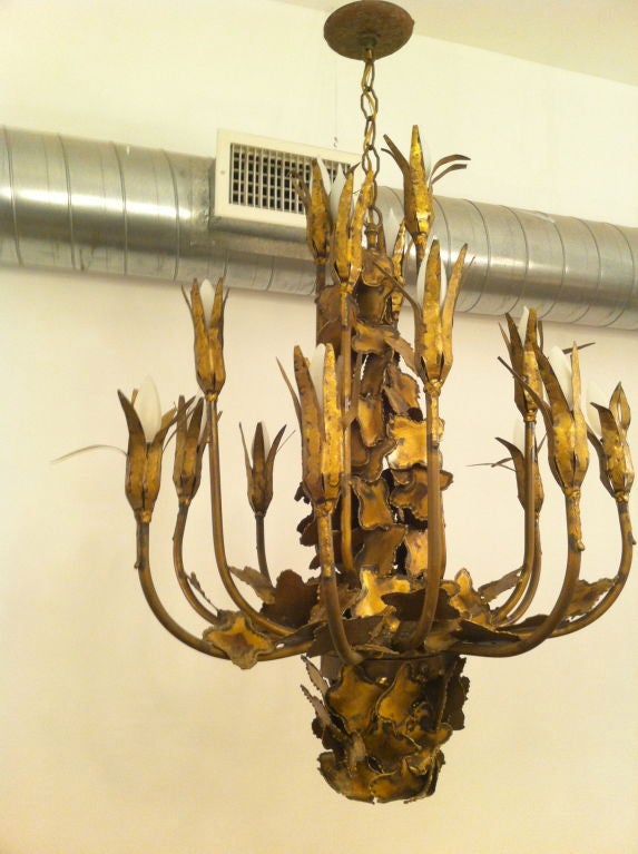 A large torch cut brutalist style floral brass chandelier by Tom Greene. It has tulip shaped sockets and had lily pad shapes decorating the body.17 light sources. One downward light near the base.