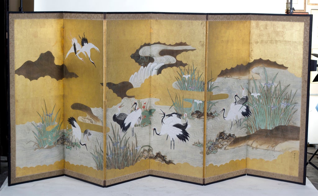 A large Japanese Edo period painted six-panel screen featuring cranes flying in gold clouds and standing in a beautiful pond. Artist signed with and old red seal. In ink, tempera paint with gilding on paper. This comes from a museum collection.