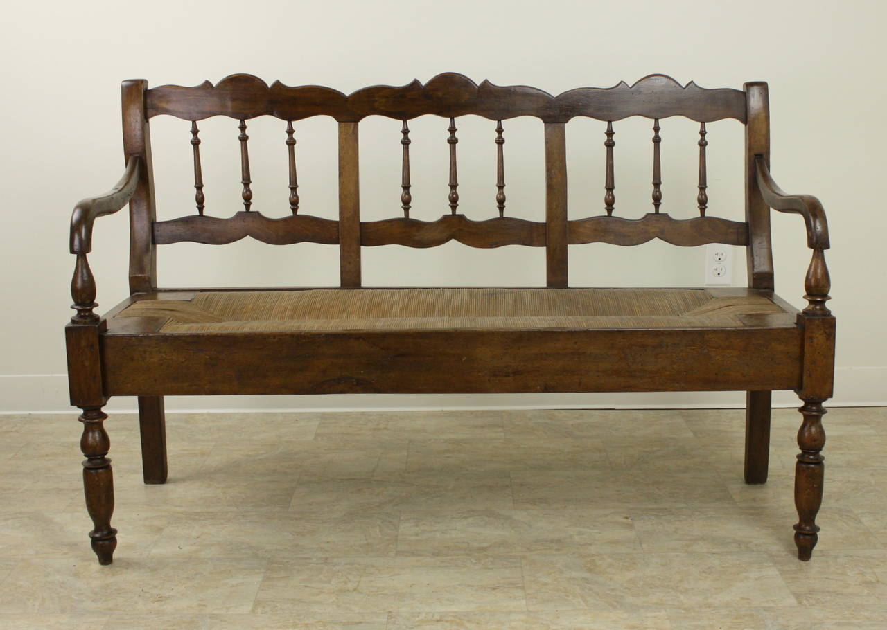Very pretty.  An antique rush seat bench from France. Note the shaped rails and spindles, curved arms, and turned legs. The chestnut has a warm color and nice patina. Seat in excellent condition.  Quite sturdy.