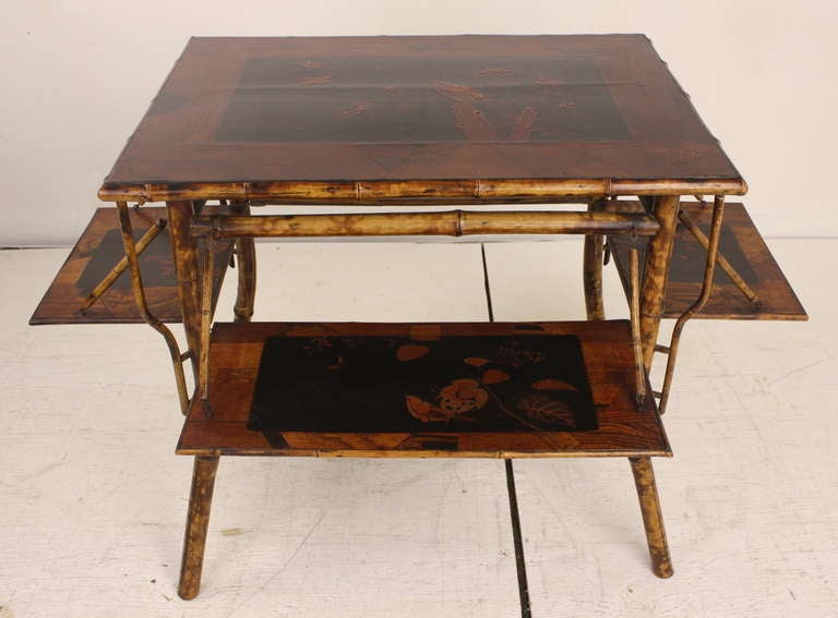 Unique English Victorian antique bamboo supper table. An intricate wood inlay surrounds the lacquer paintings on the top and the four shelves. They fold down when not in use (as seen in image 3). When they are open, the table measures 42