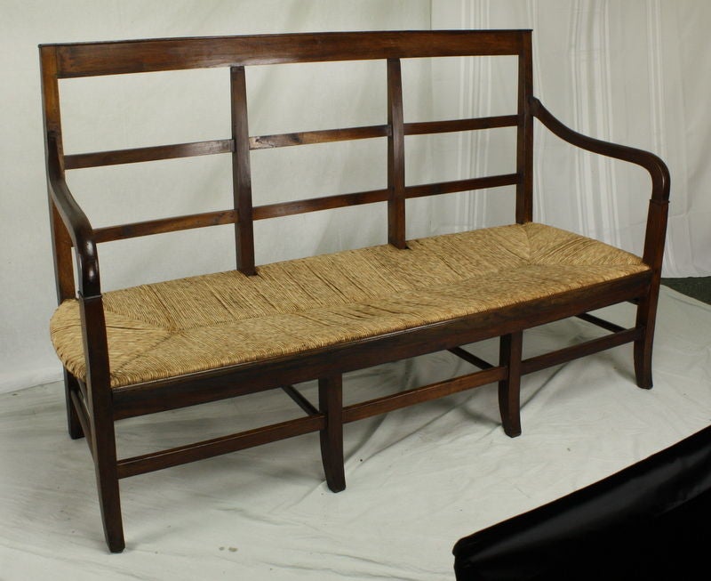 An antique rush seated bench from France. Made of walnut in a very pretty color. The high, open back and curved arms are simple and elegant. With a seat height of 15