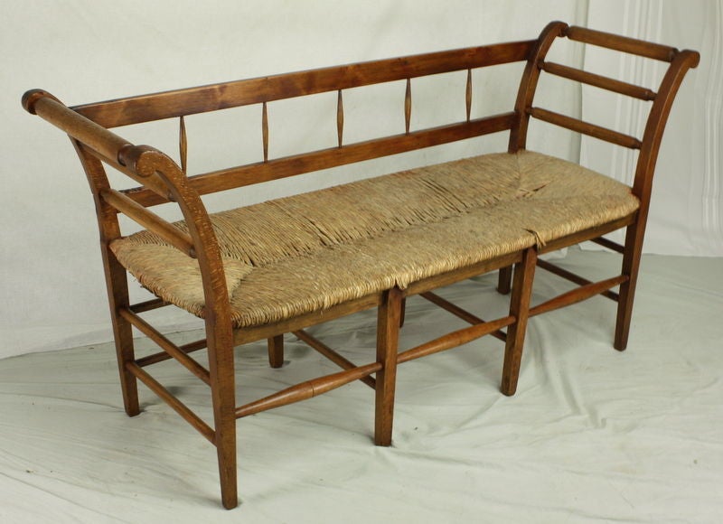 A small French rush-seated bench in honey-toned fruitwood.  The  smaller size makes this just right for the hallway or bedroom. A pretty silhouette with graceful curved arms and simple spindles.  The rush is in good condition, a few stray bits need