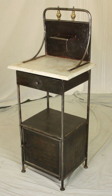 A charming French antique steel small cupboard. Very good for a  bedside cabinet.  High back gallery with small candle shelf, and is decorated with brass accents.  The marble top has an ogee edge and interesting light wear. Good height for a lamp