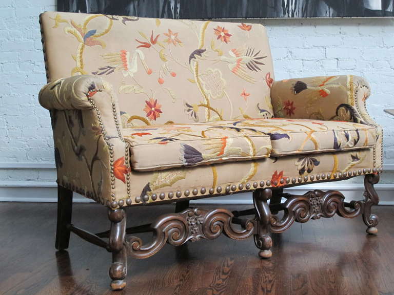 Early 20th Century Jacobean Revival loveseat covered in faded later crewel upholstery. Upholstery has fading and other wear, carved walnut base has wear and marks to the original finish.