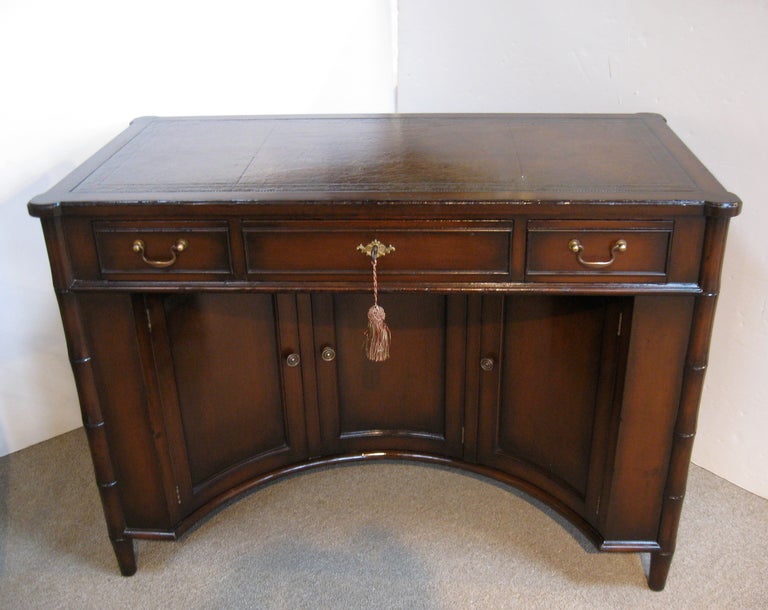 Solid wood desk with tooled leather top.  This compact desk has a recessed curved front with three doors that open to reveal additional shelves and storage.  Desk is finished on all sides, with brass hardware and faux bamboo details.
Wood has been