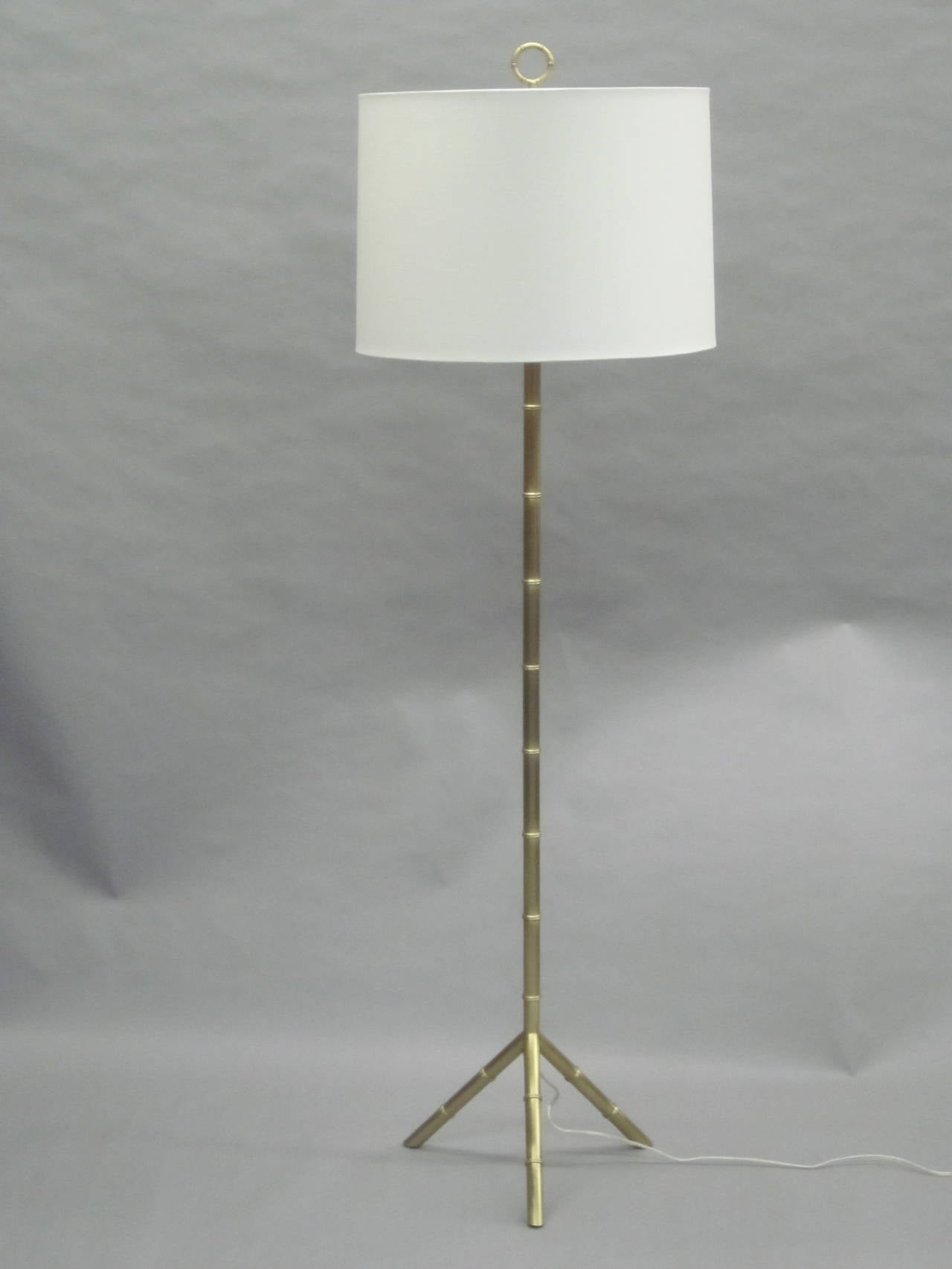 Pair of French Mid-Century style brass faux bamboo standing lamps based on models by Jacques Adnet and Maison Bagues. Each resting on a brass tripod base and having a brass stem that contains one Edison socket rated up to 150 watts, cotton shade and