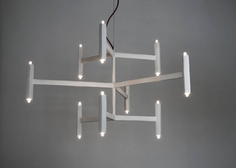 This nine-branch chandelier with a plaster finish hangs from a steel cable and seems to float in the space. From every point of view, the symmetrical design shifts perspectives. A silk electrical wire is used to add a touch of color. The fixture