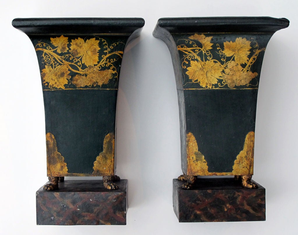 A good-scaled pair of French Empire style dark green painted tole urns with gilt decoration; each with everted lip above a tapering square body raised on paw feet resting on a square plinth; adorned with a meandering gilt grape vine perimeter band