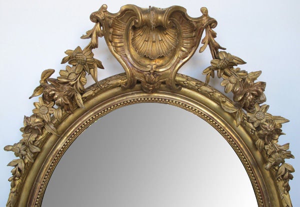 A finely-carved French Napoleon III oval giltwood mirror with shell crest and floral garland; surmounted by a bold shell crest flanked by floral festoons; all above an oval-form coved frame with beaded slip and laurel leaf outer border; surrounding