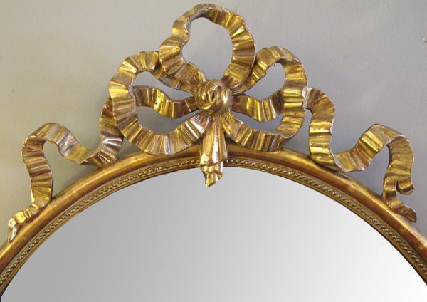 An elegant French Napoleon III oval giltwood mirror with exuberant ribbon crest; surmounted by a well-carved giltwood bow above an oval frame with beaded slip surrounding the original beveled mirror plate.
