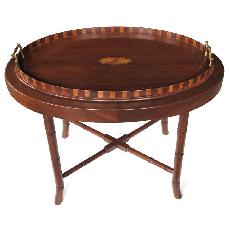 A warmly-patinated English oval mahogany inlaid tray with brass handles. The oval tray centering an inlaid fan. All within a raised gallery of alternating woods fitted with brass handles. Raised on a later faux bamboo stand.