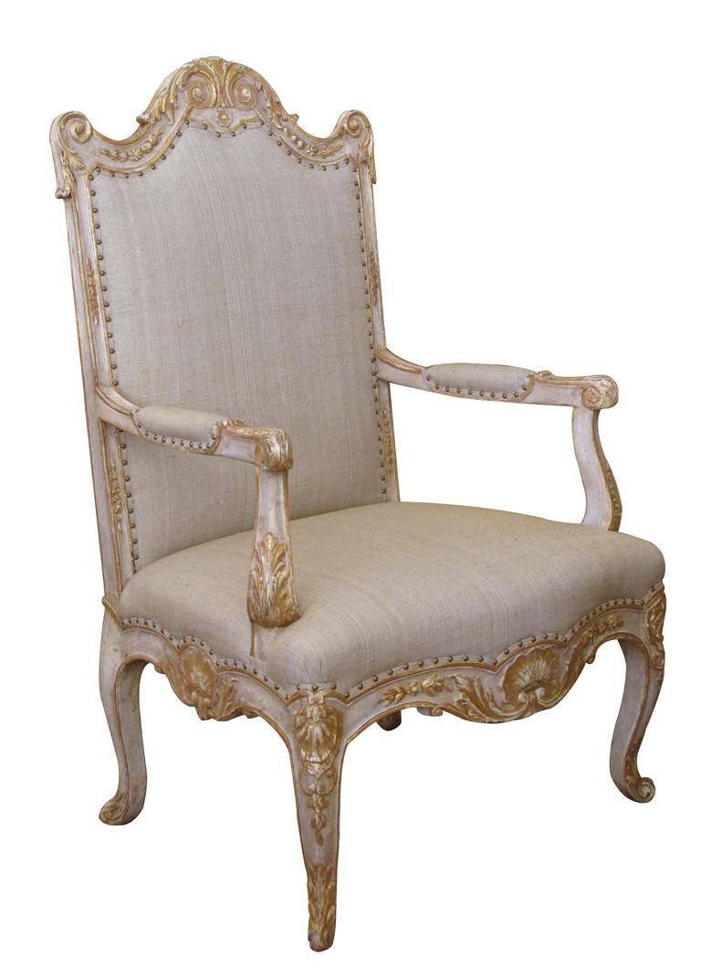A baronial French regence style ivory painted and parcel-gilt open armchair; the tall back with dramatic arching crest above a serpentine seat flanked by padded arms all above a scalloped apron raised on cabriole supports