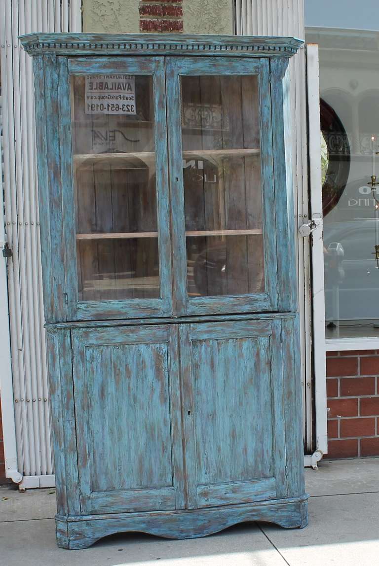 This two-piece, blue corner cupboard shows a distressed painted exterior and a white washed painted interior.  The blue painted exterior is a over a white painted undercoat.  There are two glass doors over the two-blind door base. The backing is in