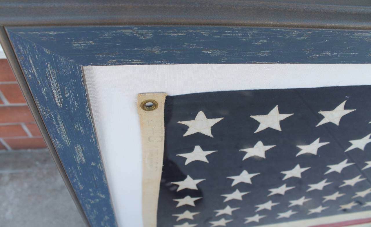 This large 48 star wool ships flag is sewn on white cotton linen and behind plexy glass is in a custom distressed blue frame. The condition is very good with minor moth bites  in some areas consistent with age and use. This has hanging loops ready