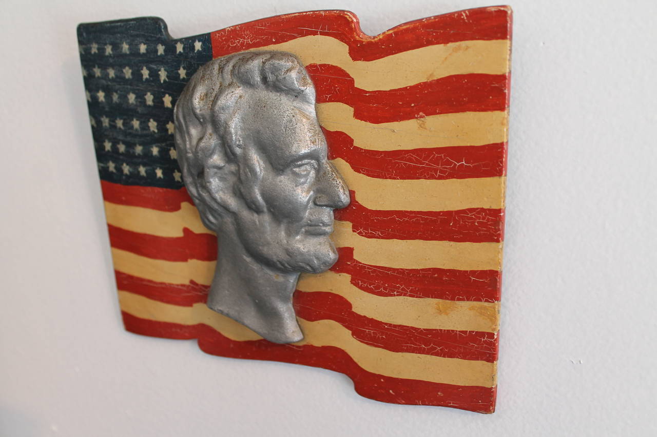 This fun and folky hand painted and carved wood flag and has a zink Lincoln head attached to the center. These were hung in the windows during patriotic events and parades. This was found in Gettysburg, Pennsylvania. The condition is mint. This is