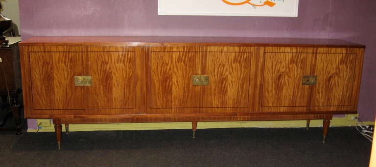 Extra long (9 ft, 2 inches) Argentine mid-century modern sideboard in highly figured satinwood. Hardware is in brass. 
This piece has been restored. Three interior cabinets. Center cabinet is mirrored and has interior lighting which will need