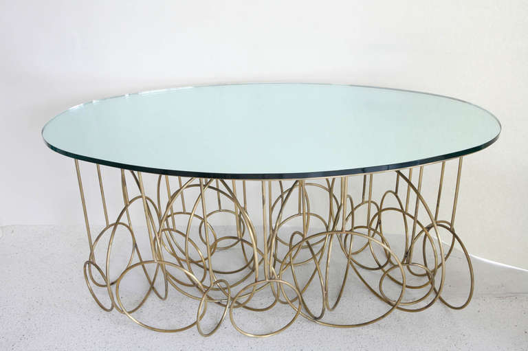 This table is constructed from solid Brass rod in a series of rings of varying  sizes forming the base of the table.  The top is mirrored glass with a blue green hue.  
This table is very much in the style of French artist Hubert le Gall.