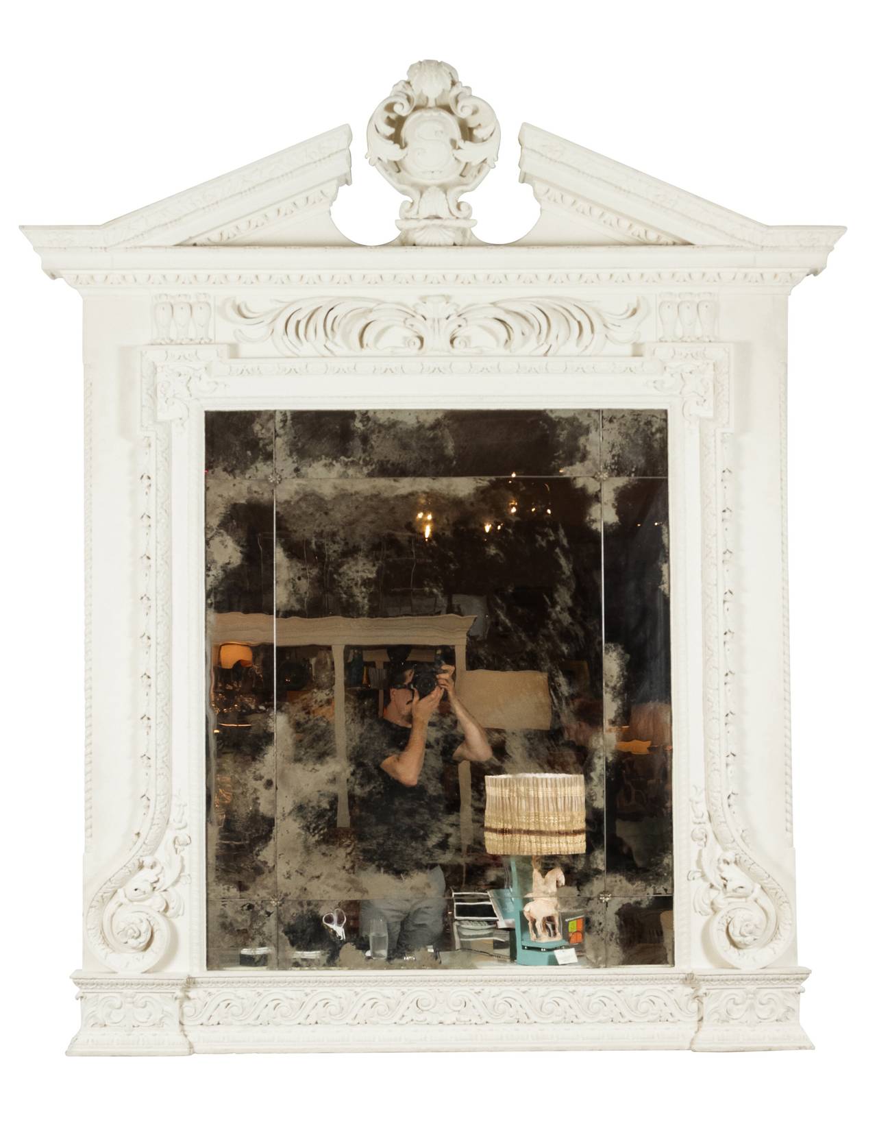 Large mirror with off-white painted frame featuring a broken pediment and scrolling motifs. Mirror has multiple, aged panels, accentuated by silver toned fleurettes. There is one main mirrored panel in the center, surrounded by eight smaller panels.