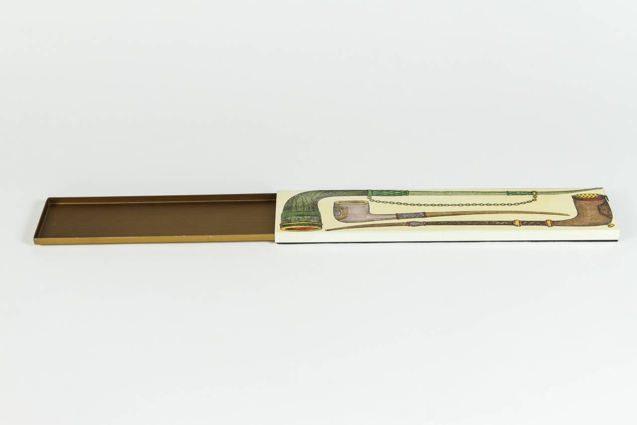 Boîte Fornasetti longue, rectangulaire, avec une charmante illustration de trois tuyaux.  Lithographie colorée à la main sur métal.  La boîte extérieure en métal coulisse sur une boîte intérieure en métal, revêtue de poudre en laiton brossé avec un