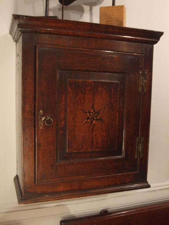 An 18th century English hanging wall cupboard in oak having swinging door on hand-forged H-hinges with holly and bog-oak inlay in the shape of a star to central door panel, molded skirting and architrave and interior shaped shelving. Outstanding