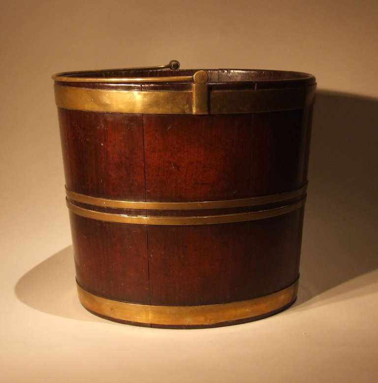 Fine George III mahogany peat bucket, having original brass bail handle over two broad and two narrow brass bands, the rare oval body in richly patinated mahogany.