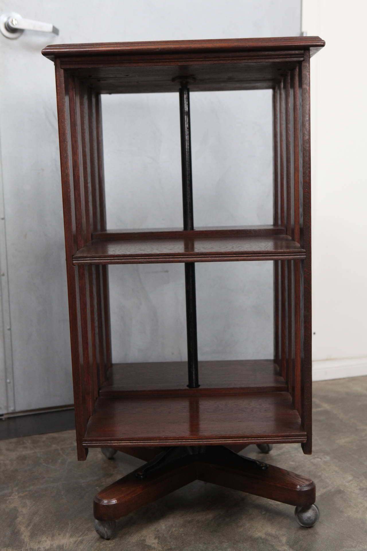 This handsome english revolving bookcase has one shelf, slated sides and a central metal column. The body is mounted on four pronged wooden feet with casters. Note the decorative brass screws and the molded edging.