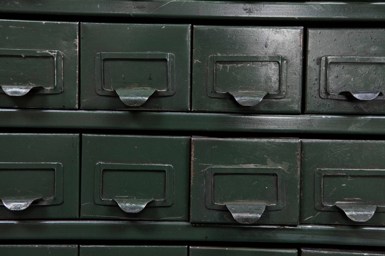 This is a great Industrial metal cabinet with legs has 100 small drawers that have label frames and pull handles. The finish on the piece has the original green and black paint with the exposed metal. The legs have been added at a later time.