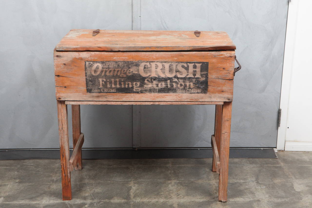 This wonderful piece was a cooler for soda pop in the early 20th century.  The cooler is made of wood, metal nails and hinges with a tin interior. The piece has very nice worn orange paint and black and white signage. The signage states: 