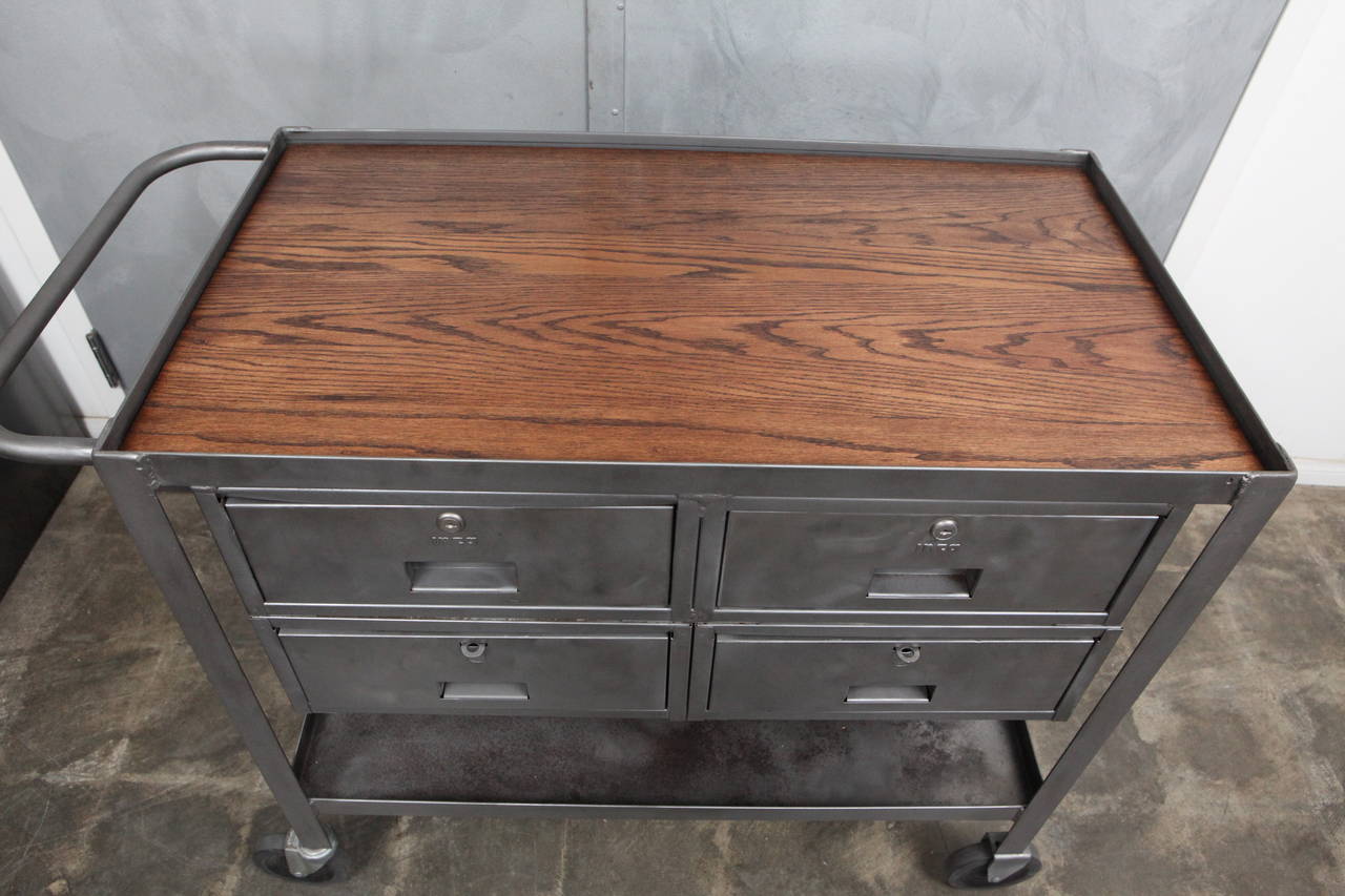 This elegantly designed polished metal Industrial rolling cart with an inset oak top has four drawers and a bottom shelf. The cart has a nicely shaped handle and four large wheels on steel casters.