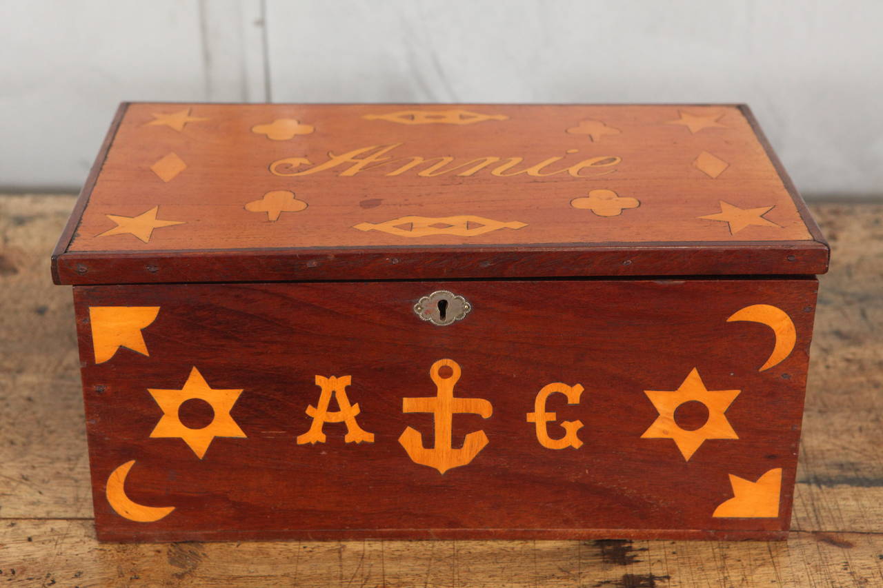 This antique sailor's box is beautifully inlaid with nautical motifs and the name of a possible sweetheart: Annie.The box has an overlapping lid and a removable compartment box lined with green felt that rests on wooden rails. There is space beneath