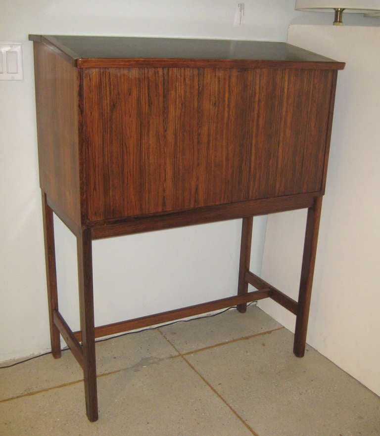 Rosewood chest on stand has slanted top for book display when closed, and opens to reveal black laminated writing/serving surface with deep well in the rear for bottles or papers. Designed for CS Mobler, this unit was offered as a combination bar