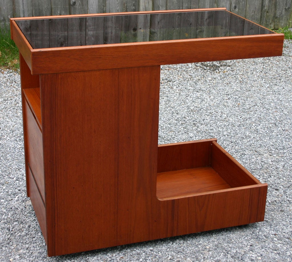 Sleek and efficient teak bar cart on metal casters; with black tinted glass top and open and closed storage.