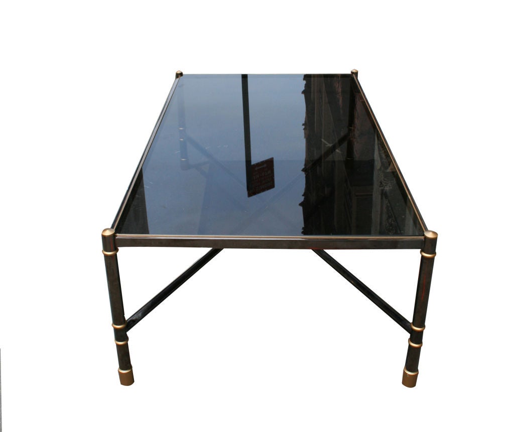 A Gunmetal and Smoked Glass Low Table by Karl Springer. USA, c. 1980s. A well proportioned low table with grey smoked glass top. Legs and cross bars in deep smoked gunmetal with matte gold accents. A campaign style low table inspired by Maison