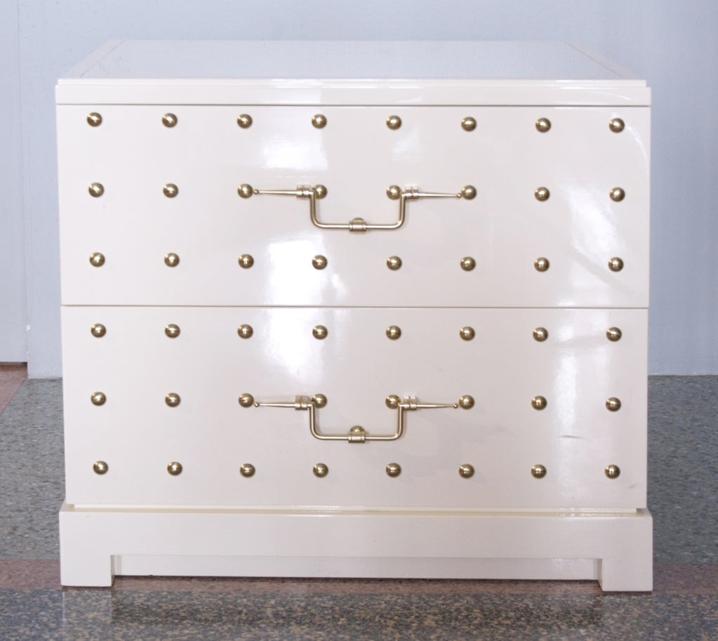 A fantastically chic pair of cream lacquered, brass studded two-drawer dressers with inset milk-glass tops by Tommi Parzinger for Parzinger Originals. In excellent restored condition these each have all original hardware, glass tops and are ready to