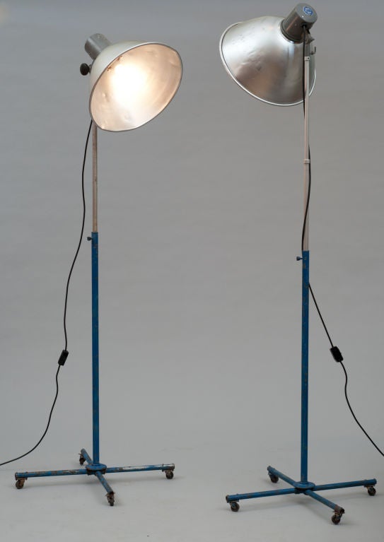 A matched pair of photographer's studio floor lamps, with adjustable head and stem.  Large spun aluminium shade with new electrical hardware for standard bulbs. X- stretcher base with 4 fully swiveling casters supports thin adjustable height stem.