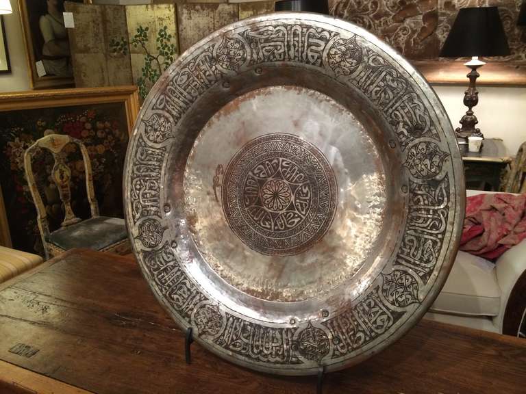 Large scale hammered and silvered copper charger decorated with etched Kufic lettering. Four wrought iron rings riveted to the reverse. With an iron stand, can also be mounted on a wall. Persian or Ottoman.