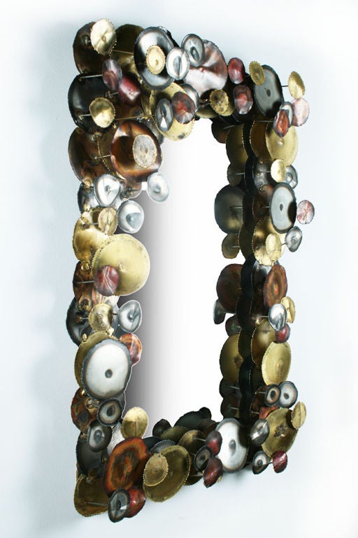 A three dimensional mirror comprised of steel, brass and bronze disks in varying sizes, varying patinas and finishes, all randomly set at different depths around a rectangular frame to create a bubbling effect.  After Curtis Jere. American, circa