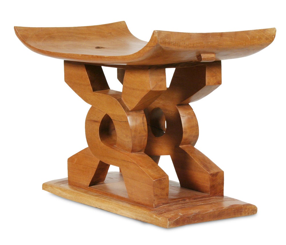 A carved wooden African stool having a curved seat raised on two carved intertwined wooden supports with a slim plinth base. Ghanaian lore explains that the central symbol carved into this cedar stool is a wisdom knot. Only the wise can untie it, so