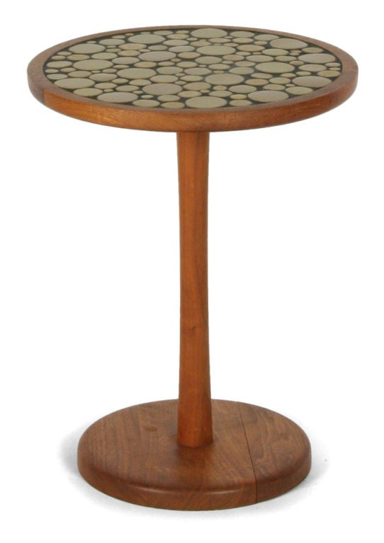 A pedestal occasional table with a round top inlaid with ochre colored circular ceramic 'coin' tiles in various diameters, the top supported by a solid turned walnut stem pedestal base. By Jane and Gordon Martz for Marshall Studios. U.S.A., circa