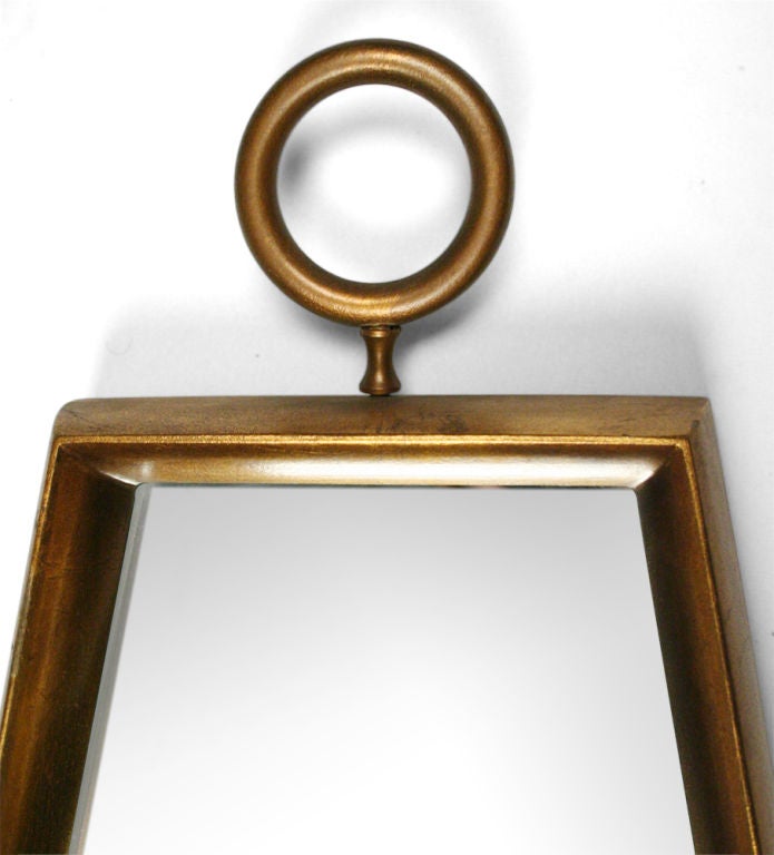 A wall-mounted mirror in a trapezoid shape with a circular 'ring' finial and slightly beveled edge, all with a gold gilded finish. After Tommi Parzinger. U.S.A., circa 1960.