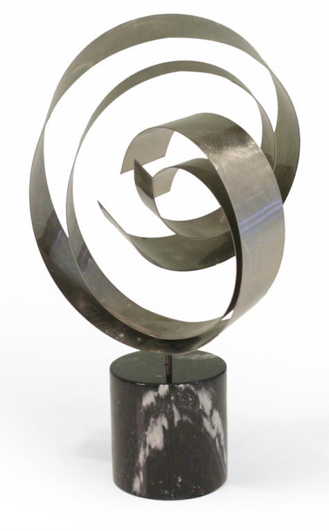 A table top sculpture with satin steel ribbon-like bands in concentric circles then mounted on a marble column base. The sculpture is designed to spin on its base to be repositioned.  By Curtis Jere. American, circa 1970.
