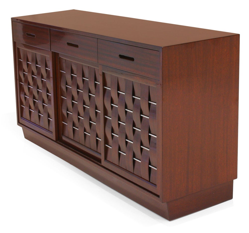 A fine cabinet comprising three drawers with interior dividers and recessed handles; each over a sliding door of walnut interwoven slats with nickeled rods, concealing three drawers on either side and an open space in the center; all raised on a