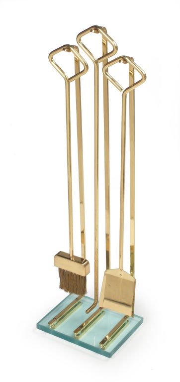 A high style set of fireplace tools comprising three tools each with a diamond form handle in all high polish brass resting on an integral brass Stand with a thick glass slab base, American, circa 1950.

