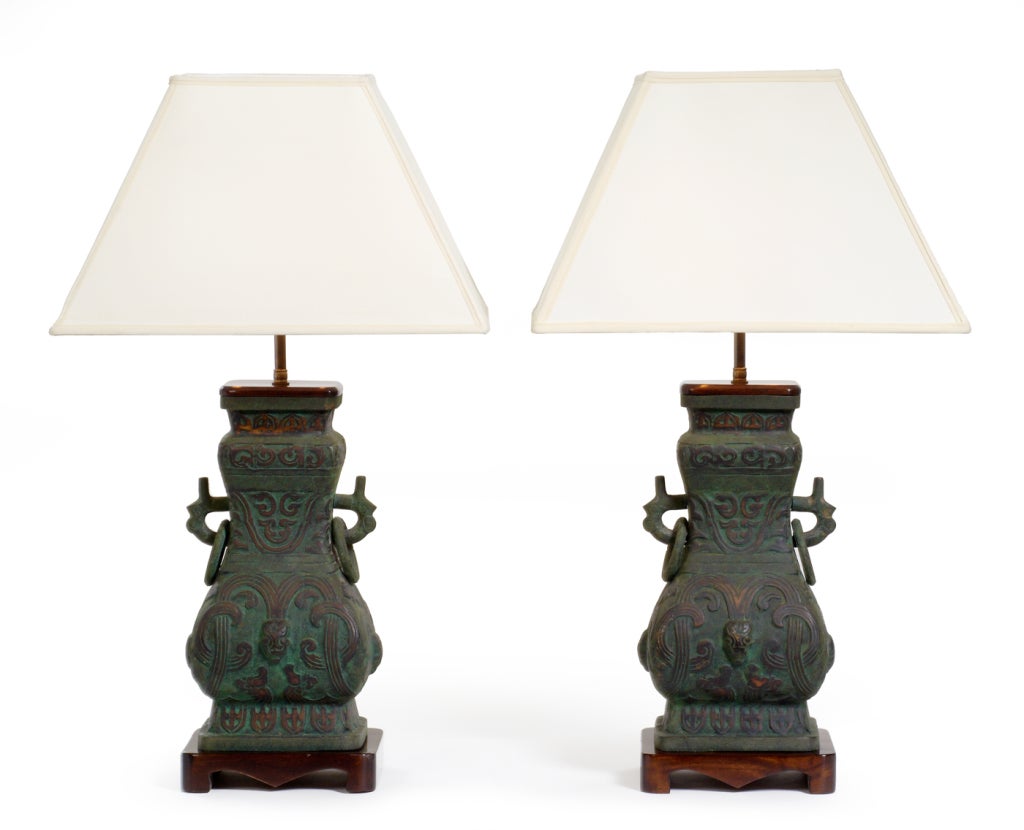 A pair of heavy table lamps in an urn shaped form of cast bronze with wood caps and mounted on carved wood bases each body has a chinoiserie design in low relief. Italian, circa 1950.