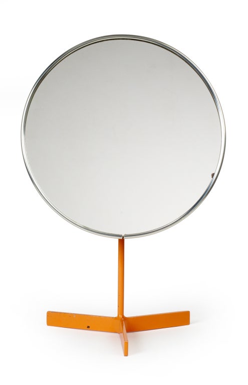 A vanity mirror in a spare Minimalist design comprising a nickeled steel thin circular frame supported on an orange lacquered tripod pedestal base. This design has been attributed to both Robert Welch and Owen F Thomas. With paper label 