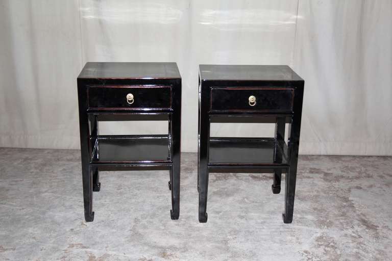 A pair of very simple and refined black lacquer end tables, each with a drawer and a shelf, brass ring pulls.