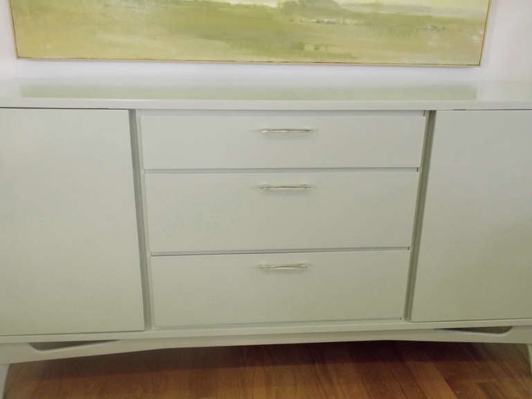 A Mid-Century sideboard/credenza. Refurbished in a celadon lacquered finish. Two opening side doors with shelves in the middle (one). Three pull-out drawers in center all with original matte nickel pulls.