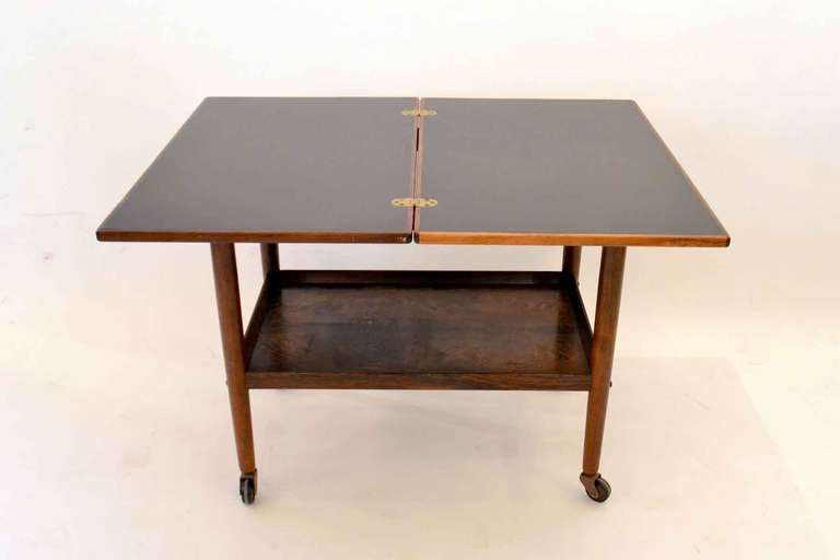 THis is a wonderful folding top bar cart  designed by Grete Jalk. The table was originally sold at Ilums Bolighus in Denmark. The table's top is made of black formica and rosewood trim. The top rotates and folds open for extension. THere is a