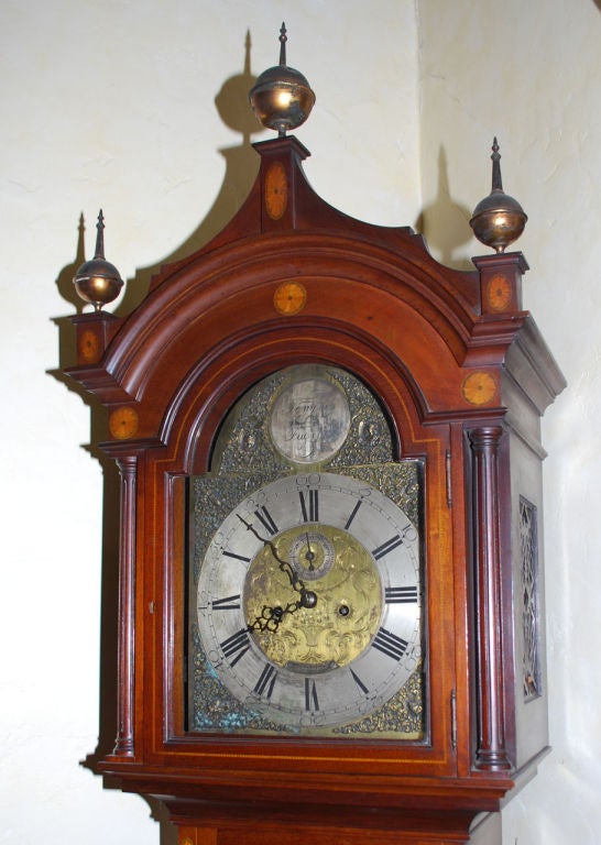 James Weir tall case clock with arched and molded cornice and brass ball finials. The brass face having cast bronze spandrels and enameled silver dials. The hood, standard and plinth inlaid with satinwood marquetry scallop roundels and stringing.<br