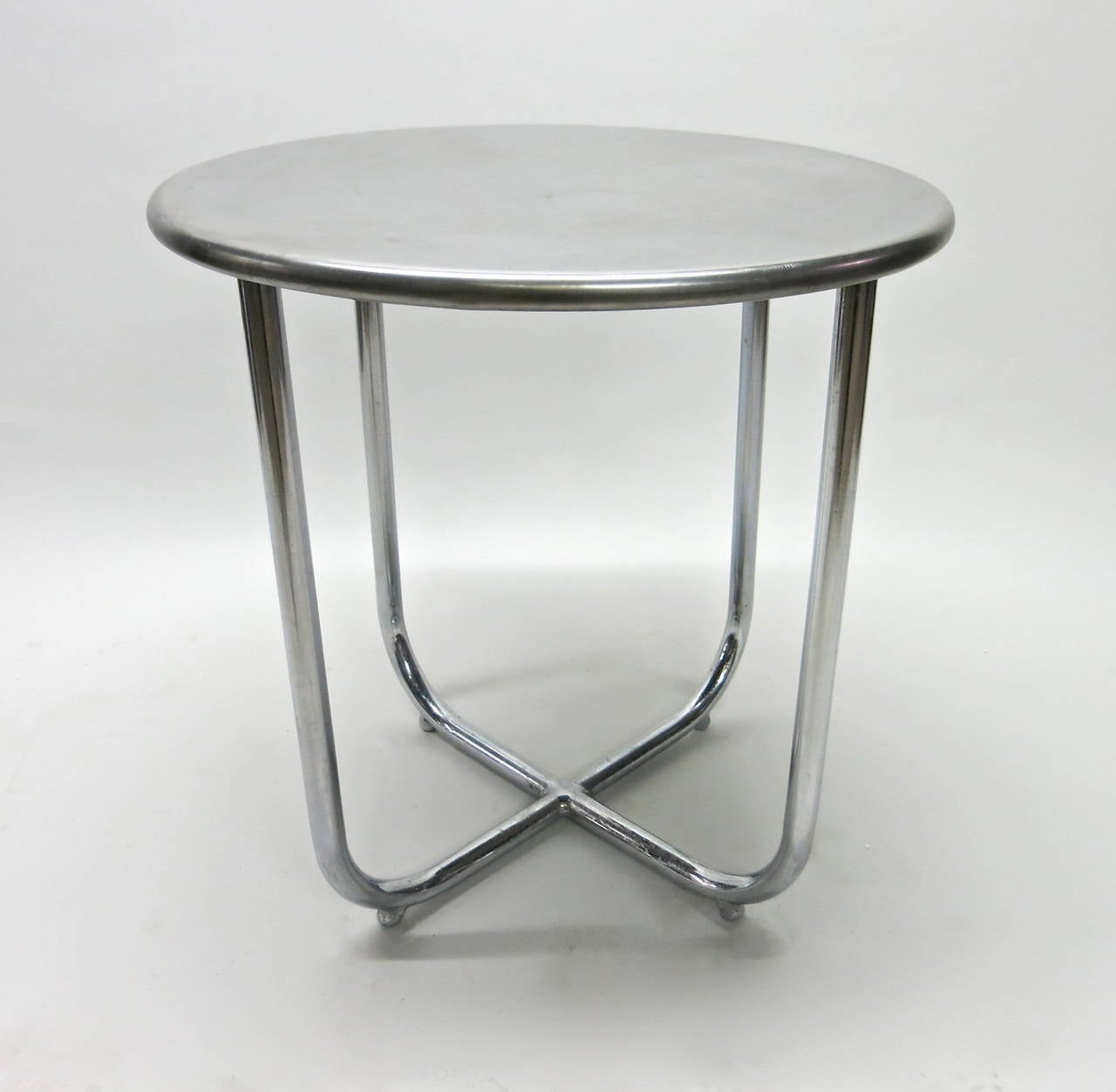 Table with a brushed steel top with curved edges supported by four tubular steel legs that bend at the bottom and cross in the center. Each leg has small foot positioned at the end of the curve.